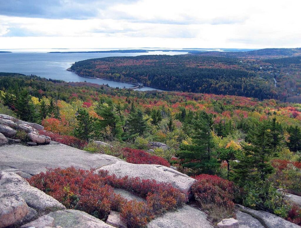 A panoramic view from Acadia National Park showing a lush, colorful autumn landscape with a lake and forest stretching to the ocean, under a cloudy sky.