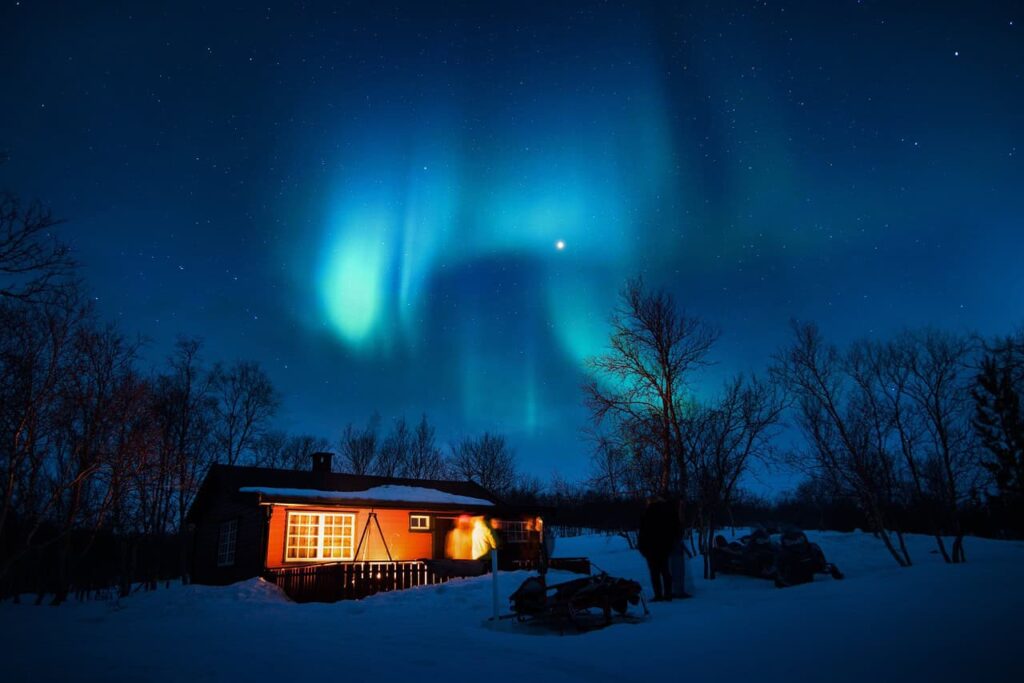 Stunning Northern Lights illuminating the sky above a cozy, snow-covered cabin in California, surrounded by silhouettes of bare winter trees.