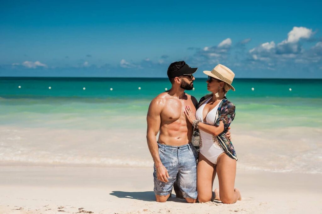 Romantic couple on a sunny Caribbean beach, standing close together in the sand, with the ocean's vivid blue water stretching out behind them.