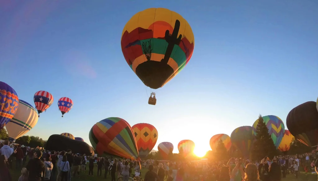 Colorful hot air balloons lifting off at sunrise during a lively balloon festival in Boise, Idaho, with a crowd of spectators gathered below.