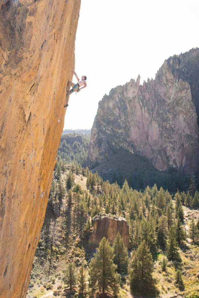 A rock climber scaling a steep orange cliff at Smith Rock State Park in Bend, Oregon, surrounded by a stunning landscape of rugged mountains and green forests.