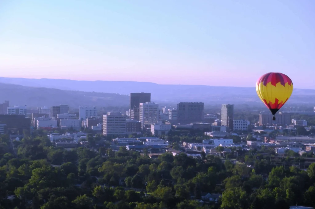 A colorful hot air balloon rising above Boise, Idaho, with a panoramic view of the city's skyline and surrounding hills at sunrise."