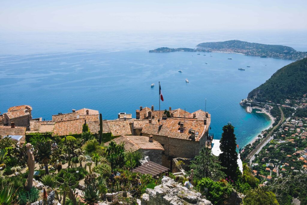 Panoramic view of Eze, France, featuring charming stone buildings with terracotta rooftops surrounded by lush, exotic gardens. The French flag waves in the breeze, and the clear blue waters of the Mediterranean Sea stretch out in the background, dotted with yachts and framed by the lush coastline.