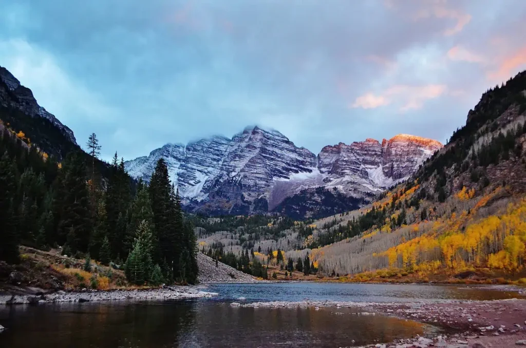 A stunning view of snow-capped mountains with a tranquil lake in the foreground, surrounded by pine trees and autumn foliage in vibrant colors showing the valley view hot springs