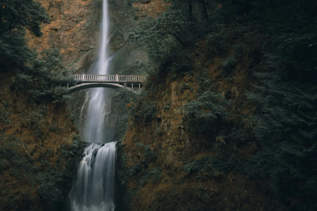 A majestic waterfall cascading under a charming arched pedestrian bridge surrounded by dense greenery in Oregon.