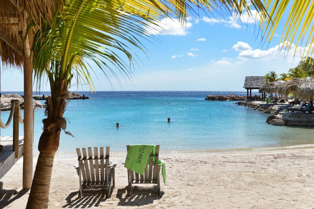 Tranquil beach in Curacao with clear turquoise water, lounge chairs shaded by palm leaves, and a wooden pier extending into the ocean