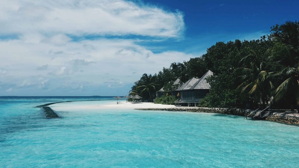 Turquoise waters and white sandy beach lined with thatched-roof bungalows in the Maldives under a bright blue sky.