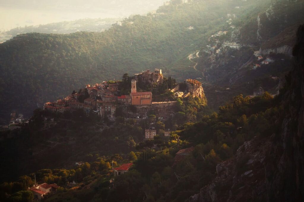 A breathtaking aerial view of the medieval village of Eze, France, bathed in golden light at sunset, surrounded by lush, forested hills and rugged mountain terrain, creating a serene and mystical atmosphere