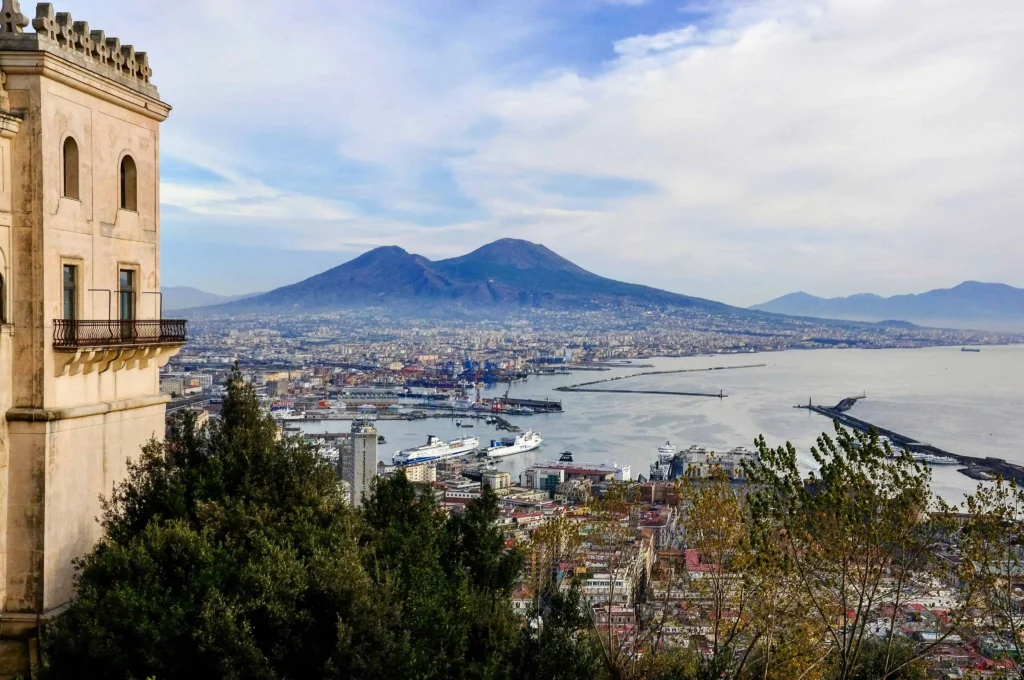 A panoramic view of Naples, Italy, from a hilltop, showcasing the harbor, cityscape, and Mount Vesuvius on a partly cloudy day.