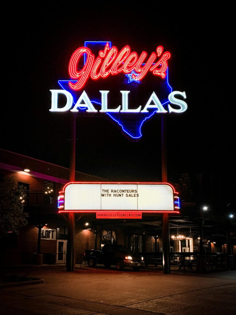 Gilley’s Dallas neon sign glowing brightly at night, with a marquee announcing a performance by The Raconteurs with Hunt Sales, set against the dark sky.