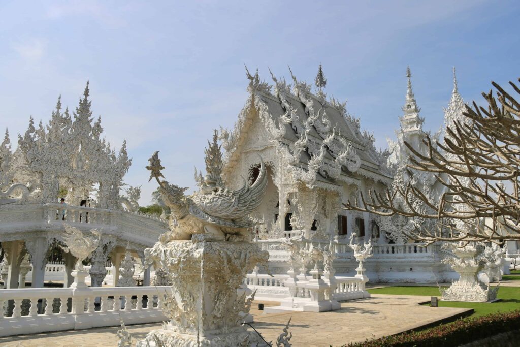 Wide-angle view of Wat Rong Khun, the White Temple in Thailand, featuring ornate, intricate carvings and a serene backdrop under a soft blue sky.