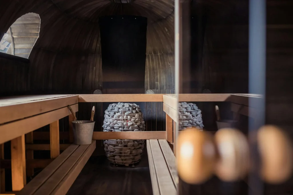 Interior of a cozy wooden sauna with benches, a bucket, and a modern stone heater in the center, creating a warm and relaxing ambiance