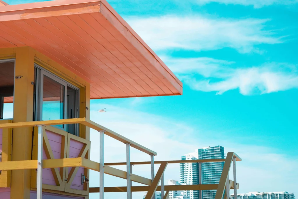 Vibrant lifeguard tower with pink and yellow accents against a bright blue sky, with modern city buildings visible in the background