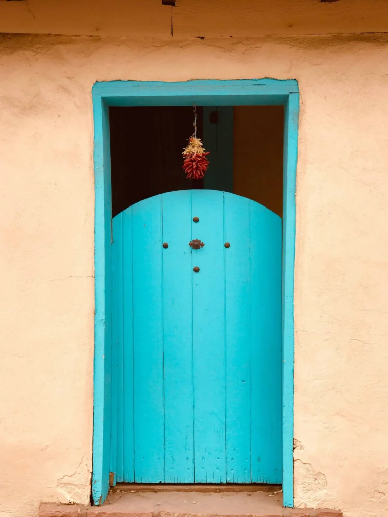 A vibrant turquoise door adorned with hanging red chilies, a quintessential feature of Santa Fe's unique Southwestern architecture