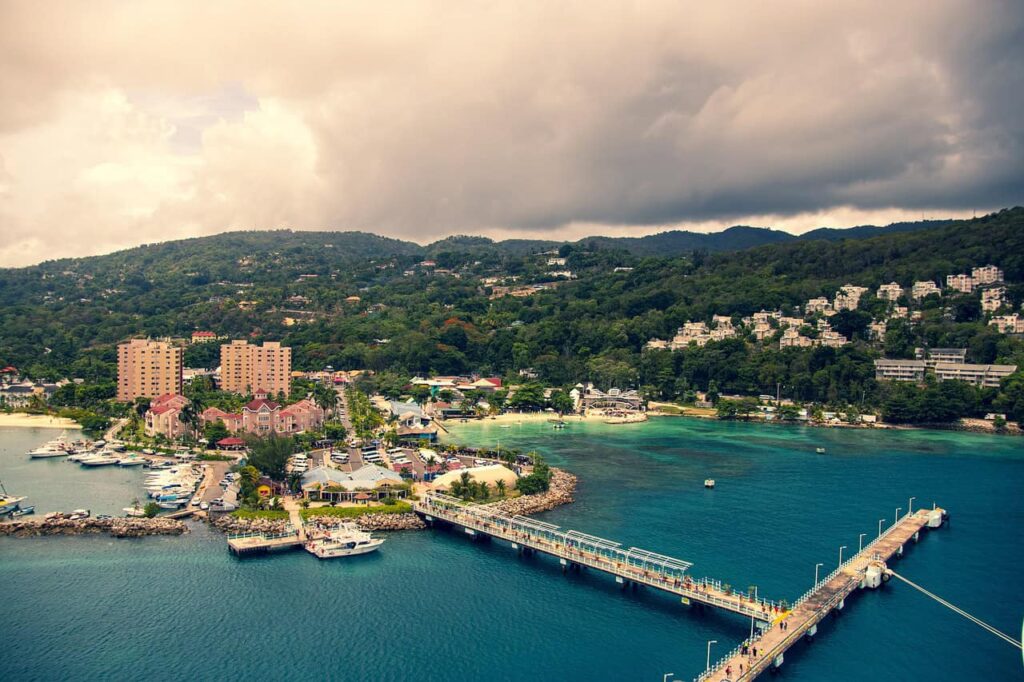 Aerial view of Ocho Rios, Jamaica, showcasing the coastline with lush green hills in the background and a bustling pier extending into the turquoise sea.