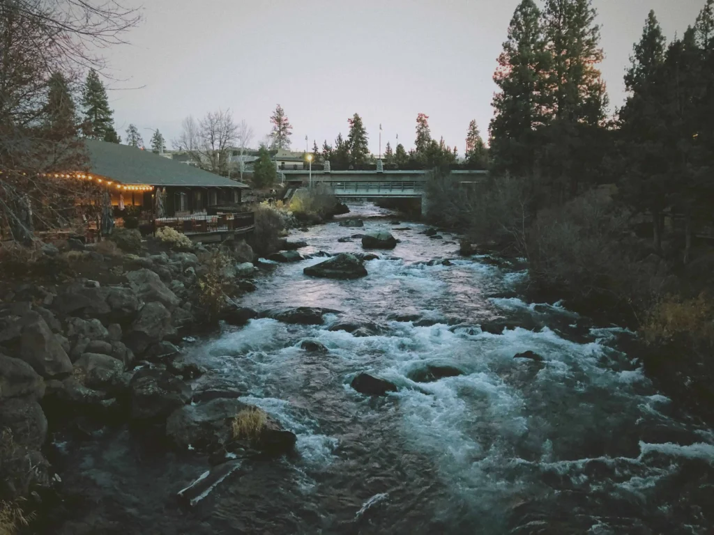 A rushing river with rocks and trees on both sides, seen from a bridge in Bend, Oregon, with warm lights glowing from nearby buildings at dusk