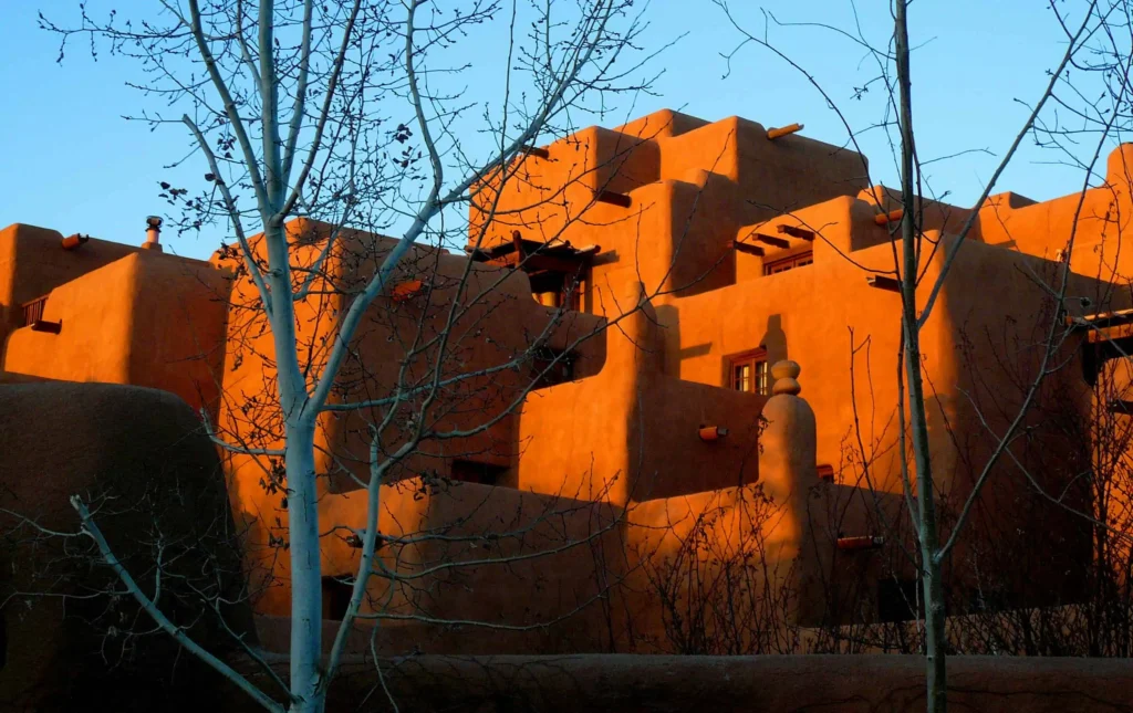 The warm, golden glow of the iconic Pueblo-style adobe buildings in Santa Fe during sunset, framed by the bare branches of winter trees.