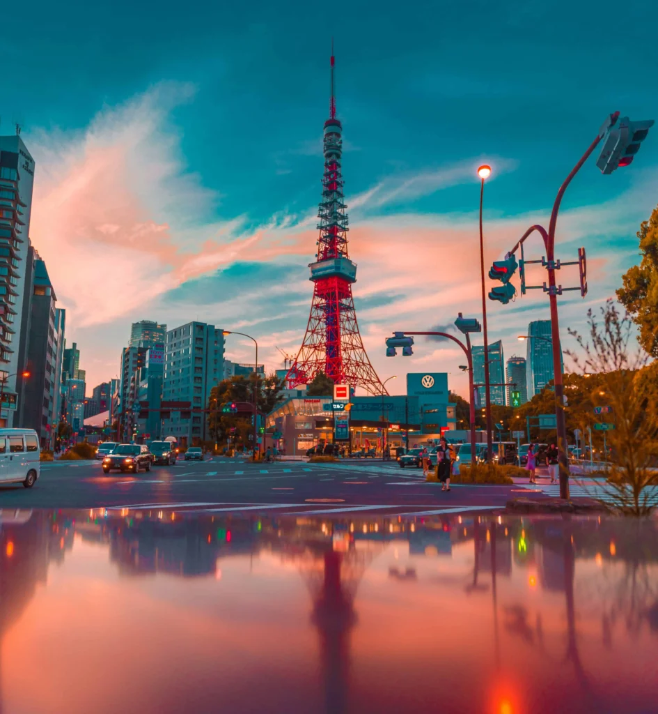 Tokyo Tower illuminated against a vibrant sunset sky, surrounded by modern city buildings and streetlights, with its reflection visible on a shiny surfac