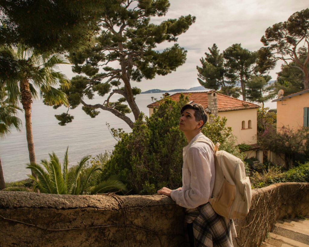 A traveler with a backpack stands on a rustic stone pathway surrounded by lush greenery, palm trees, and scenic Mediterranean views, enjoying a moment of tranquility in a charming coastal village."