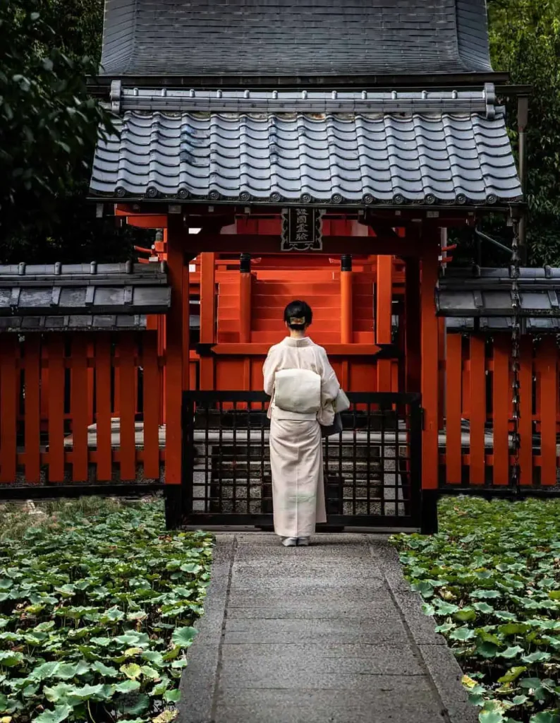 A woman dressed in a traditional Japanese kimono standing in front of a vibrant red shrine gate, surrounded by lush greenery and serene atmosphere.