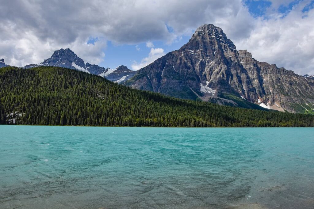 A picturesque view of Rocky Mountain National Park featuring a turquoise alpine lake in the foreground, dense green pine forests, and towering rugged mountain peaks under a partly cloudy sky.