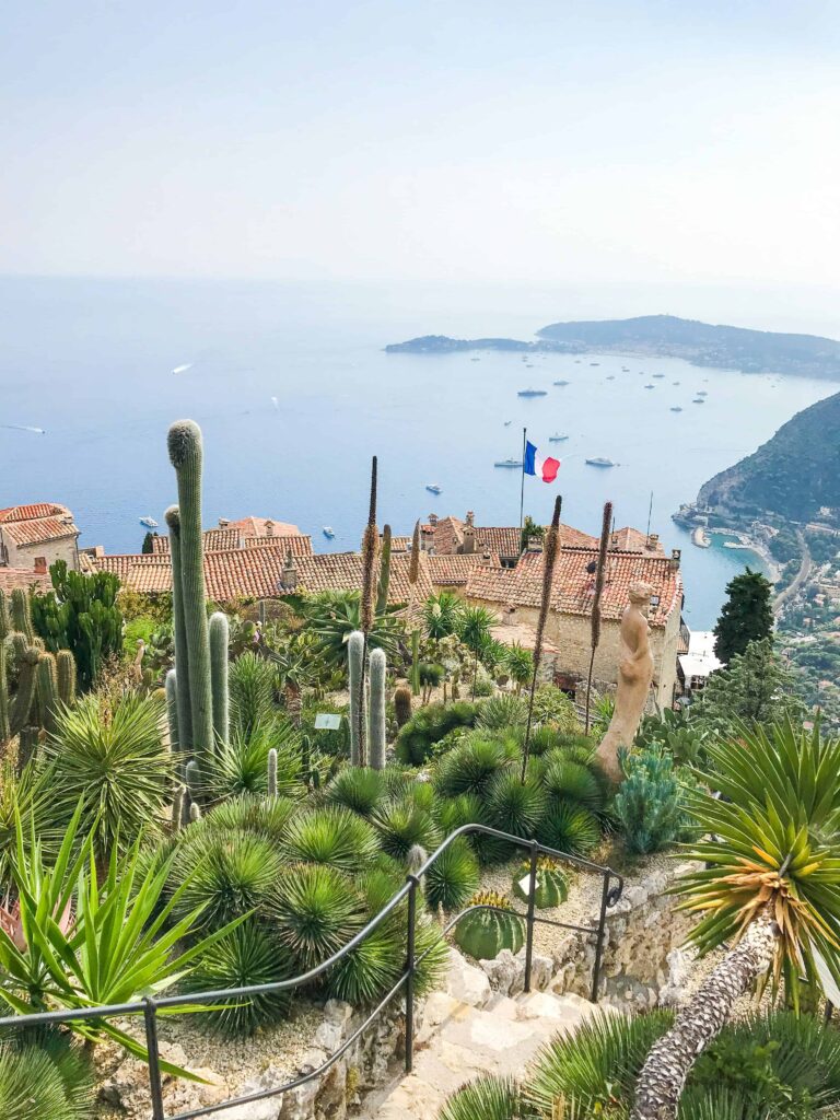 View from the Exotic Garden in Eze, France, showcasing a lush collection of various cacti and succulents against a backdrop of stone buildings with terracotta rooftops. The French flag waves prominently, and the shimmering Mediterranean Sea stretches out to the horizon, dotted with boats and surrounded by rugged coastline