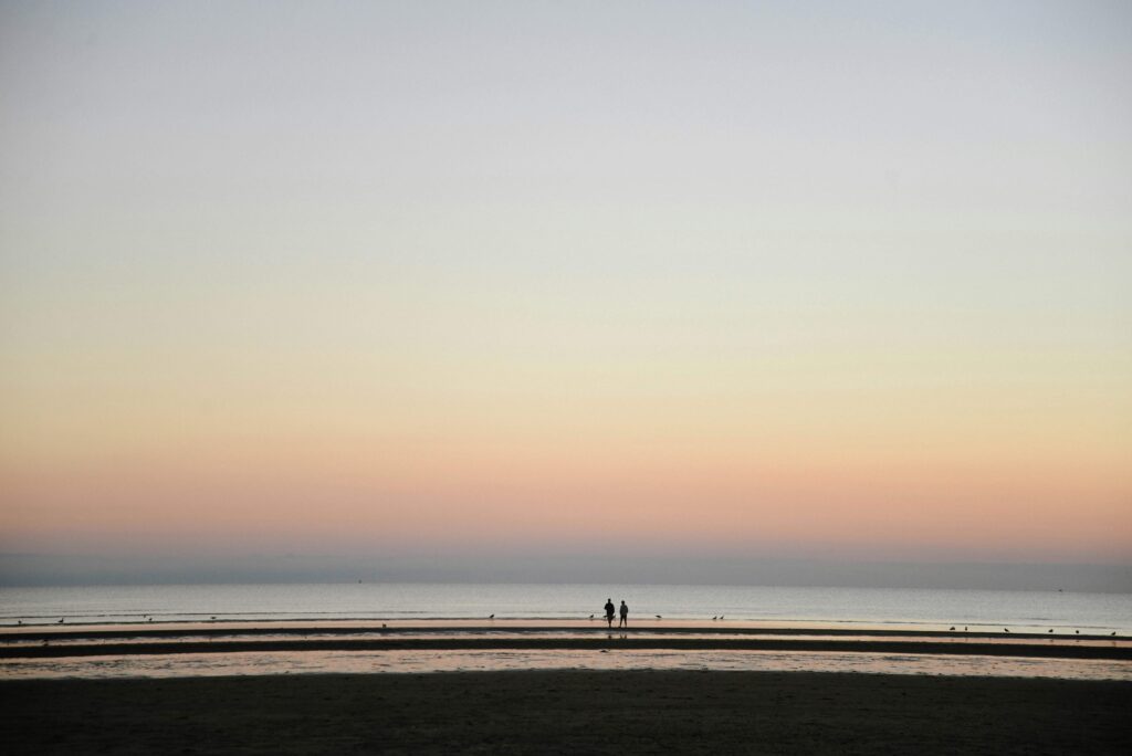 A peaceful scene of Cape Cod Bay at sunset, with soft pastel colors in the sky and two people walking along the water's edge.