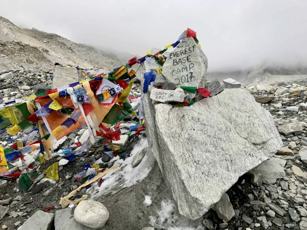 Everest Base Camp marker surrounded by colorful prayer flags, set against a misty, rocky background, commemorating climbers' journeys