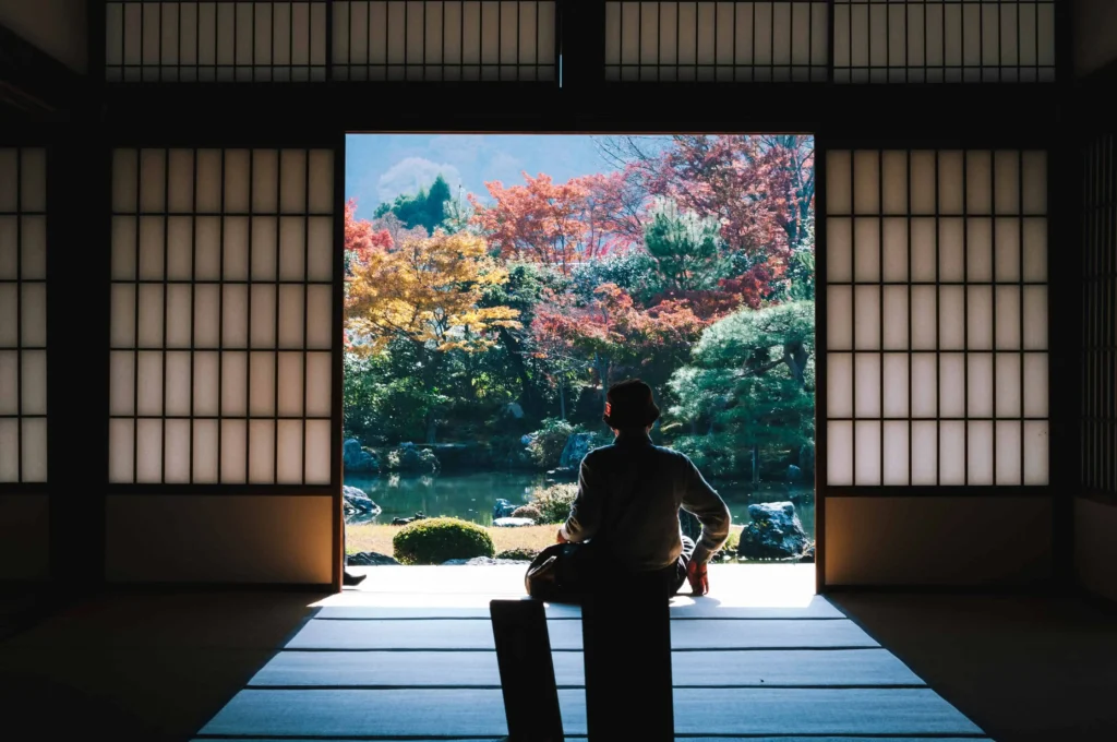 A person sitting inside a traditional Japanese temple, overlooking a serene autumn garden with vibrant foliage in Kyoto, Japan.