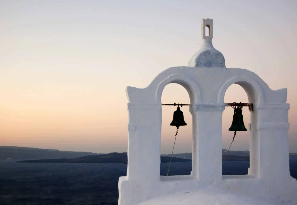 Honeymoon destinations all inclusive Greece: Iconic white church bell tower overlooking the Aegean Sea during a serene sunset in Santorini, Greece, showcasing traditional Cycladic architecture and romantic scenery.