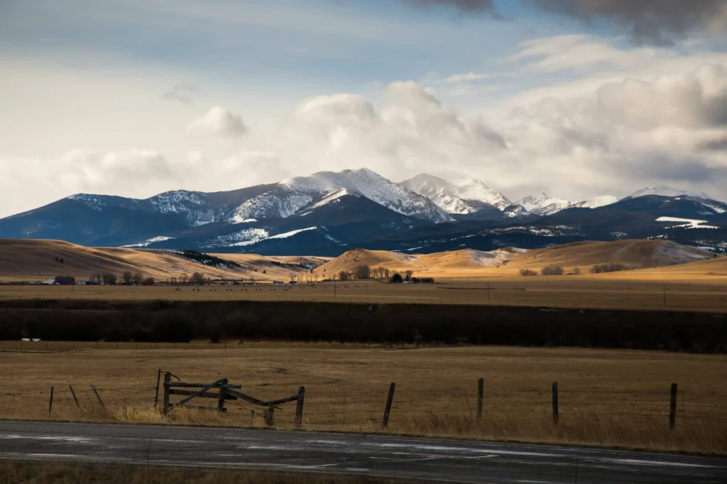 A scenic view of the Montana countryside near Bozeman, showcasing golden fields, a rustic wooden fence, and snow-capped mountains under a cloudy sky.