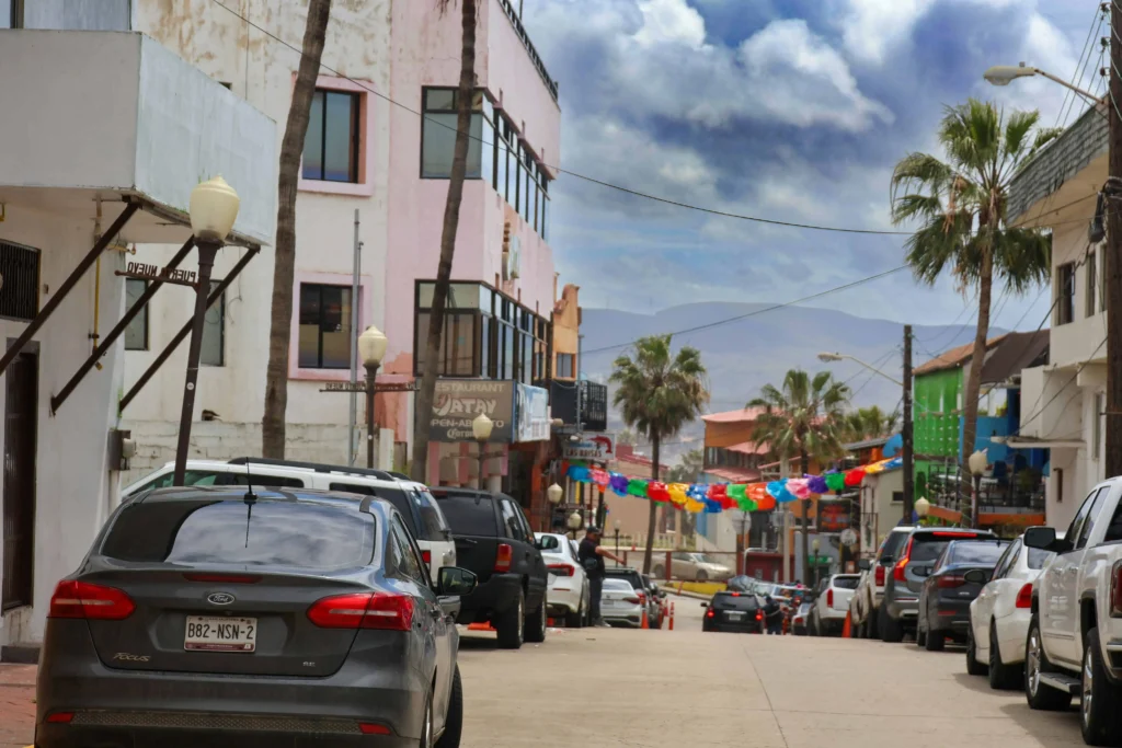 A lively street in a colorful coastal town with vibrant buildings, palm trees, and cars parked along the roadside. Festive bunting adds charm to the atmosphere.