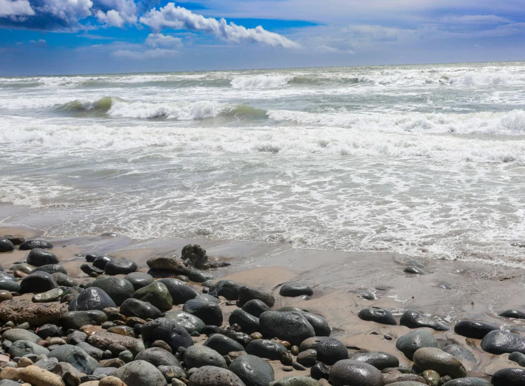 A scenic view of a rocky beach with smooth, dark stones meeting the crashing waves of the ocean under a cloudy blue sky.