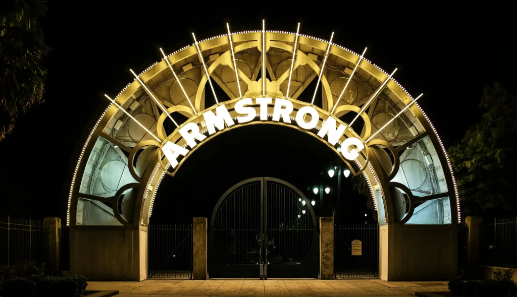 Illuminated entrance arch of Louis Armstrong Park at night, showcasing its iconic design with glowing lights and the name "Armstrong" prominently displayed.