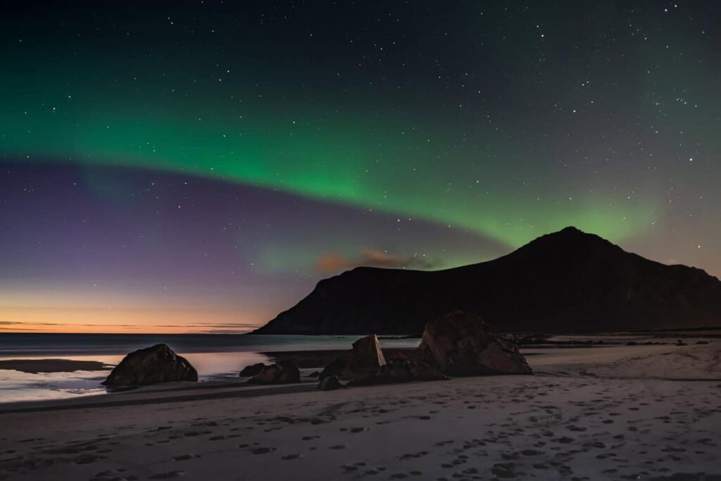 Soft green and purple Northern Lights stretching across the night sky over a serene beach with rocks and a silhouette of a mountain in the background.