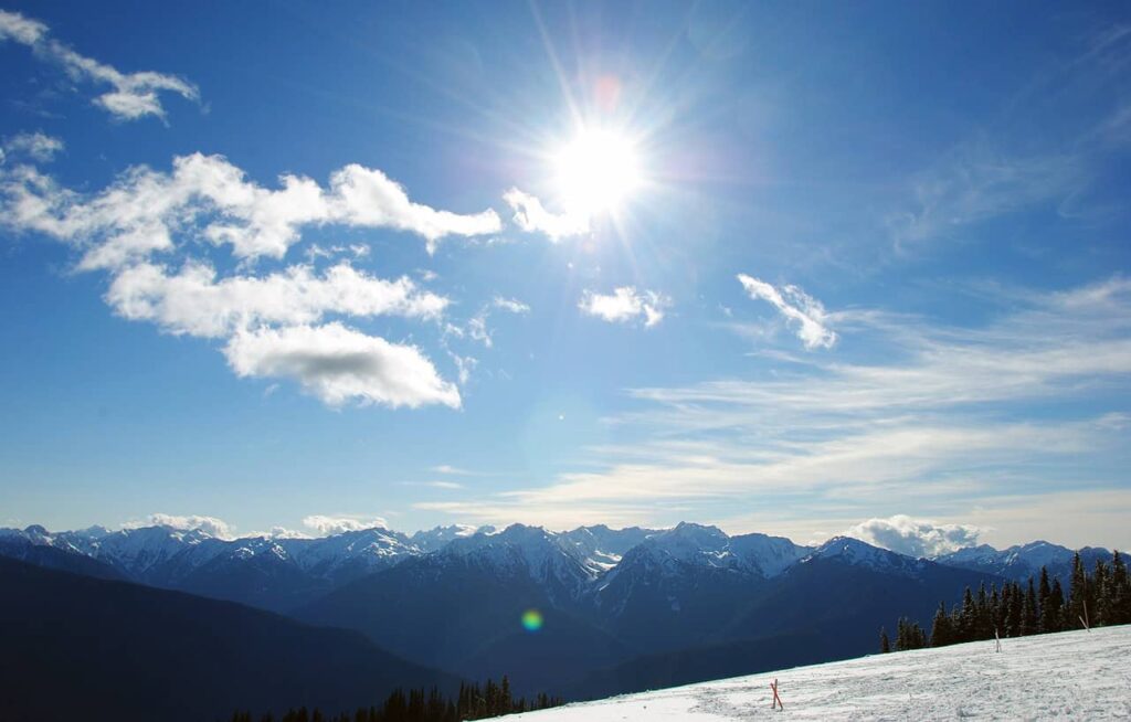 A bright, sunny day over the snow-covered peaks of Olympic National Park, showcasing the park’s stunning mountainous landscape and clear blue skies.