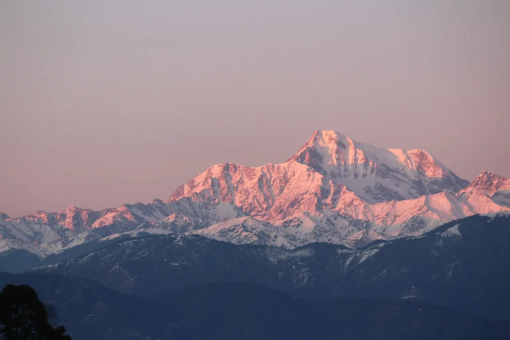 A breathtaking view of Mount Everest at sunset, with its snow-capped peak glowing in shades of pink and surrounded by the majestic Himalayan mountain range under a clear sky.