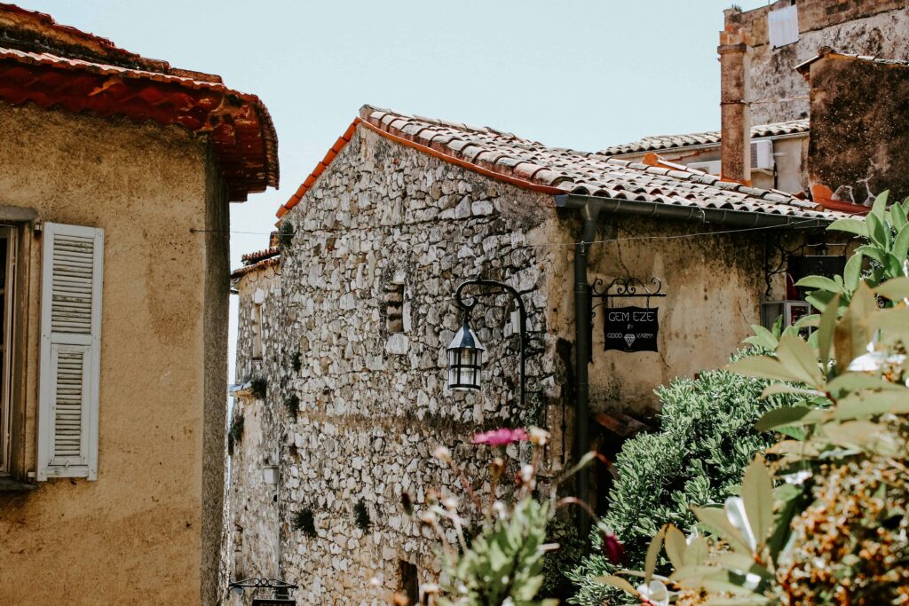 Charming stone buildings in Eze Village with rustic shutters and a quaint sign for a local gem shop, surrounded by blooming flowers and vibrant greenery under a sunny sky.