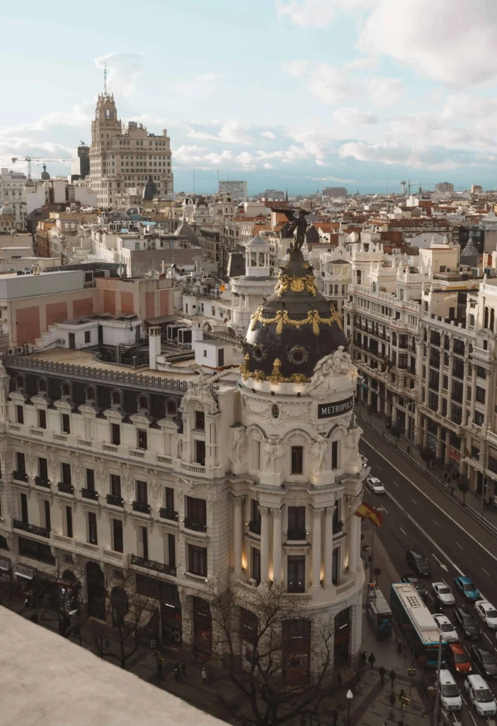 The ornate Metropolis Building in Madrid, with its gilded dome and detailed architecture, surrounded by bustling streets and a vibrant cityscape.