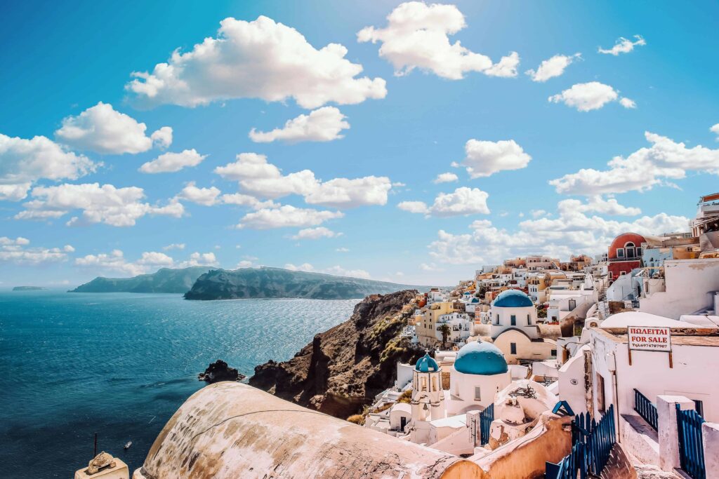 Scenic view of the beautiful Santorini coastline with iconic blue-domed buildings overlooking the shimmering Aegean Sea, under a sky filled with fluffy clouds — perfect for a romantic Greece honeymoon in 2025.