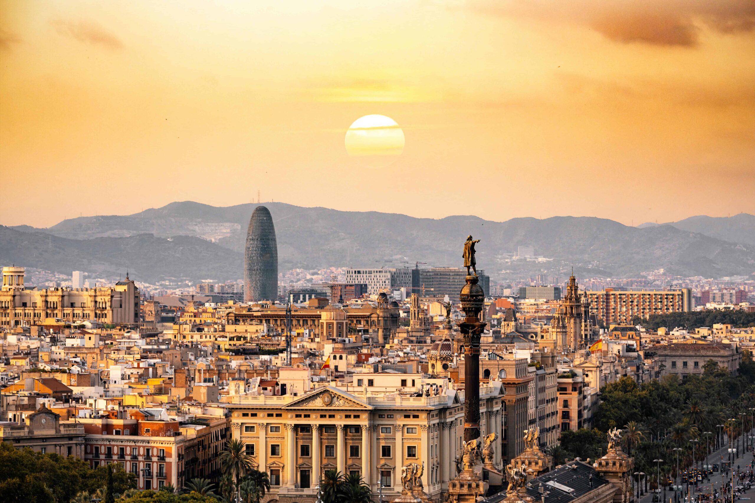 Sunset view over Barcelona, highlighting the Christopher Columbus monument and the city’s skyline, with the Torre Glòries and distant mountain range silhouetted under a warm golden sky."