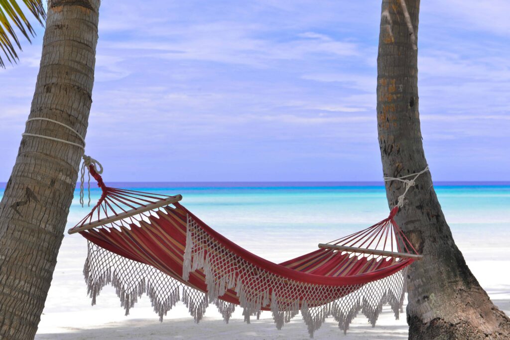 A vibrant red hammock hanging between two palm trees on a pristine Caribbean beach, symbolizing relaxation and affordability for one of the 7 cheap Caribbean vacations in 2025. The turquoise ocean stretches into the horizon, inviting a serene weekend escape.