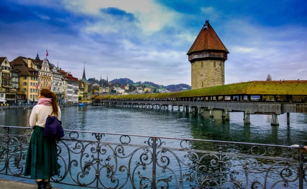 A scenic view of Lucerne, Switzerland, with a woman gazing at the historic Chapel Bridge (Kapellbrücke) and surrounding medieval architecture reflected in the calm waters of Lake Lucerne under a cloudy sky.