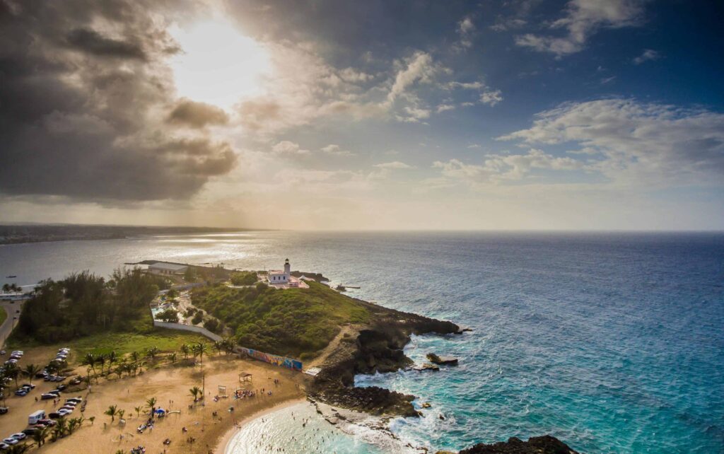 Lighthouse perched on a cliff in Puerto Rico overlooking the ocean at sunset, with golden light breaking through dramatic clouds and illuminating the shoreline below.