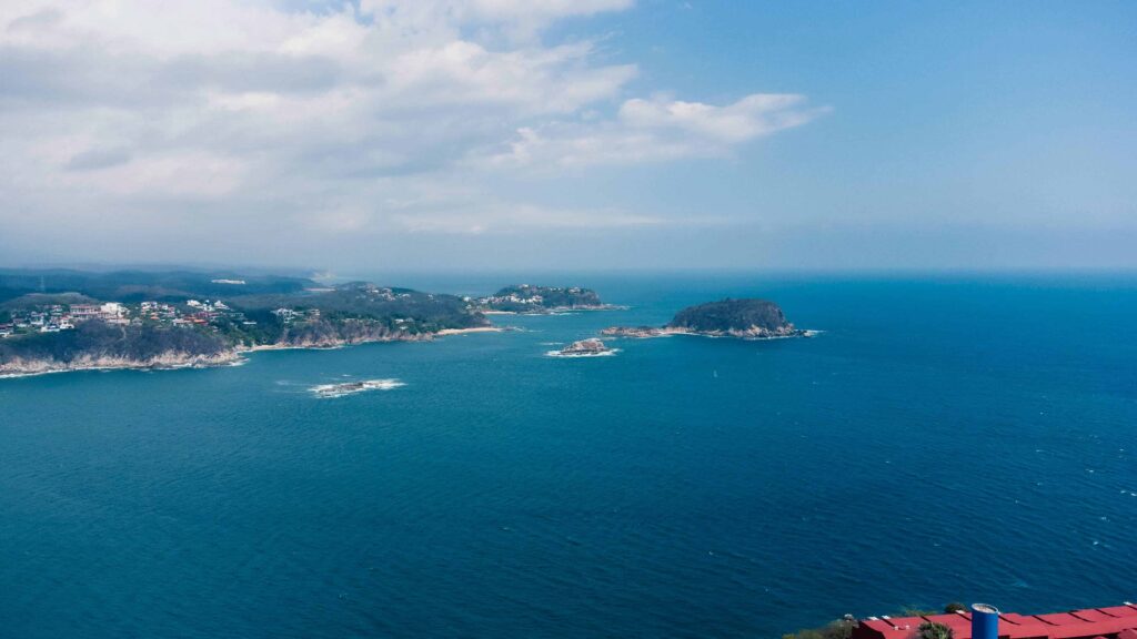 Panoramic view of Huatulco's coastline with turquoise waters and coastal cliffs under a partly cloudy sky.