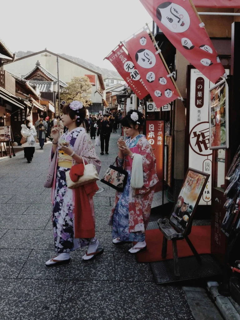 Two women wearing traditional Japanese kimonos enjoying street food while strolling through a bustling market street in Kyoto.