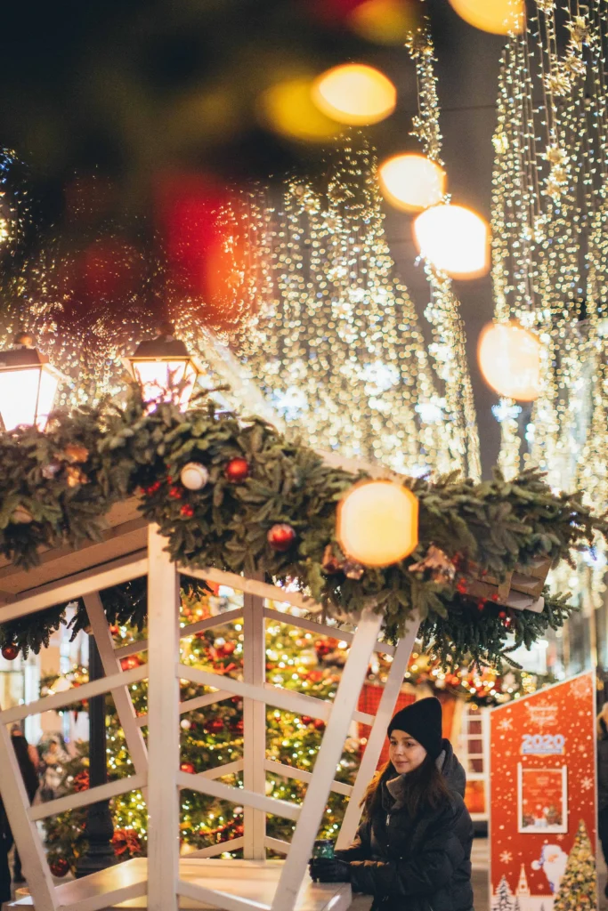 A cozy Christmas market stall adorned with festive greenery, twinkling lights, and ornaments, with a woman enjoying a warm drink in the holiday ambiance.