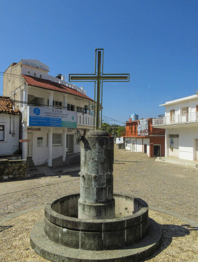 A large stone cross standing in the middle of a town square under a clear blue sky in La Crucecita, Huatulco, Mexico