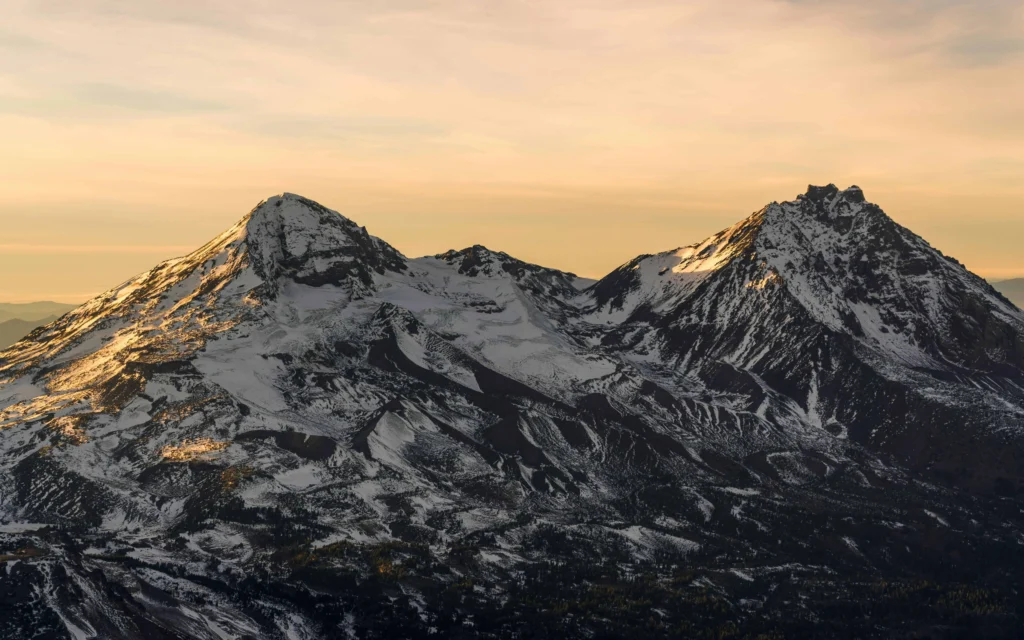 A breathtaking view of two snow-capped peaks in the Cascade Mountains during sunset, with soft golden light illuminating the rugged terrain near Bend, Oregon.