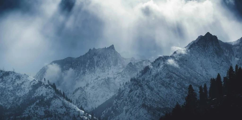 Snow-covered mountain peaks in a dramatic misty landscape with clouds casting ethereal light, highlighting the rugged beauty of the scene